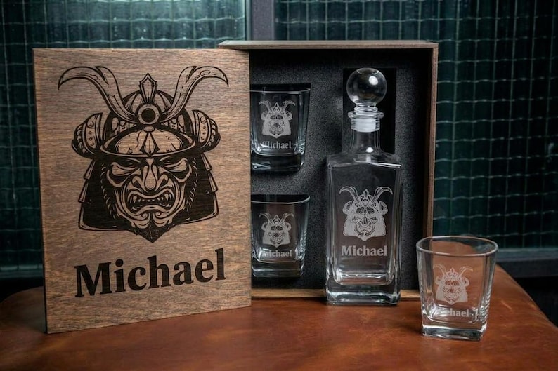 May include: A wooden gift box containing a clear glass decanter and three matching square glasses. The box lid and each glass feature an engraved samurai helmet graphic and the name Michael. The decanter has a round glass stopper.