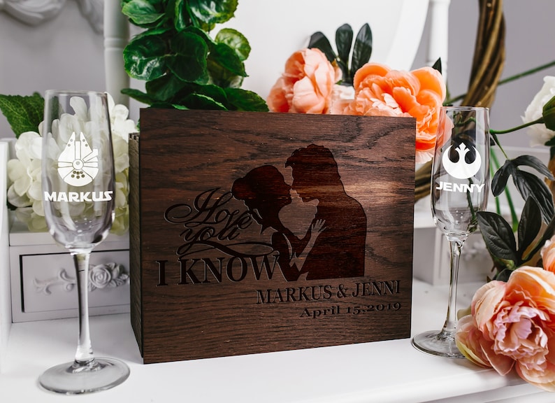 May include: A wooden box with a laser engraved design of a couple embracing. The text "I love you I KNOW" is engraved on the box. The box is decorated with two champagne flutes, one with a Star Wars Millennium Falcon design and the other with a Rebel Alliance symbol. The flutes are engraved with the names "Markus" and "Jenny". The box also has the names "Markus & Jenny" and the date "April 15, 2019" engraved on it.