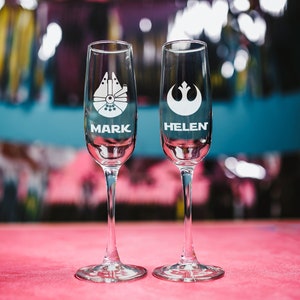 May include: Two clear glass champagne flutes with personalized names "Mark" and "Helen" etched on the bottom. The flutes are decorated with Star Wars themed images, a Millennium Falcon and the Rebel Alliance symbol.