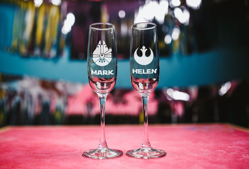 May include: Two clear glass champagne flutes with personalized names "Mark" and "Helen" etched on the bottom. The flutes are decorated with Star Wars themed images, a Millennium Falcon and the Rebel Alliance symbol.