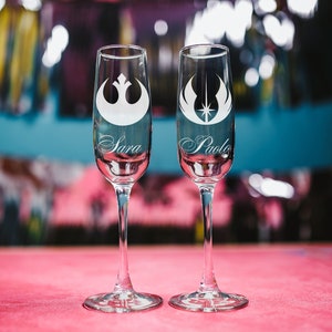 May include: Two champagne flutes with etched Star Wars Rebel Alliance symbols and the names "Sara" and "Paolo" on a pink surface.