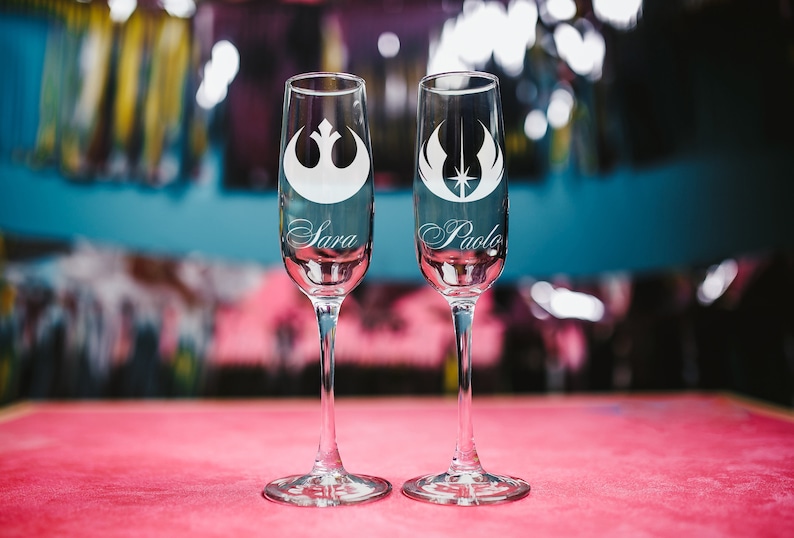 May include: Two champagne flutes with etched Star Wars Rebel Alliance symbols and the names "Sara" and "Paolo" on a pink surface.