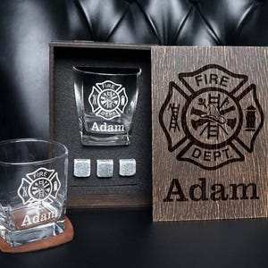 May include: A set of two clear glass tumblers, three grey stone whiskey cubes, and a dark wooden box. Each glass features an etched fire department emblem and the name Adam. The wooden box lid is engraved with the same emblem and the name Adam.