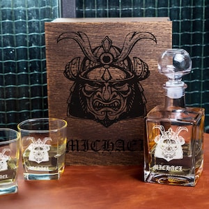 May include: A clear glass decanter and two matching square glasses, each etched with a samurai helmet design and the name MICHAEL. A dark wooden box stands behind them, featuring a large, detailed black engraving of a samurai helmet.