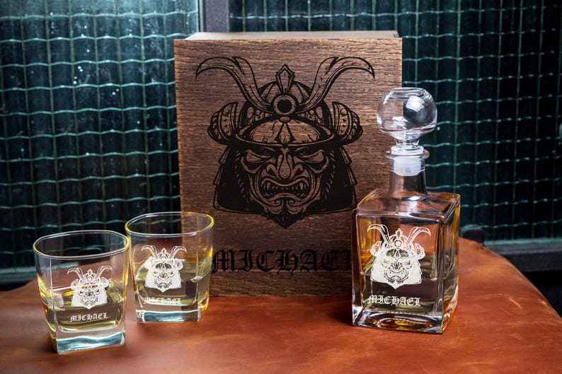 May include: A clear glass decanter and two matching square glasses, each etched with a samurai helmet design and the name MICHAEL. A dark wooden box stands behind them, featuring a large, detailed black engraving of a samurai helmet.