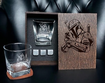 May include: A whiskey glass set featuring two clear glasses with etched Mandalorian helmet designs, three grey stone ice cubes, and a dark wooden box with a matching laser-engraved Mandalorian helmet graphic on the sliding lid.
