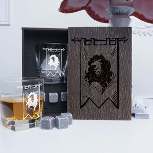 May include: Two clear glass whiskey tumblers with a white lion emblem on a banner design. The tumblers are in a wooden box with a matching wooden panel featuring the same lion emblem design.