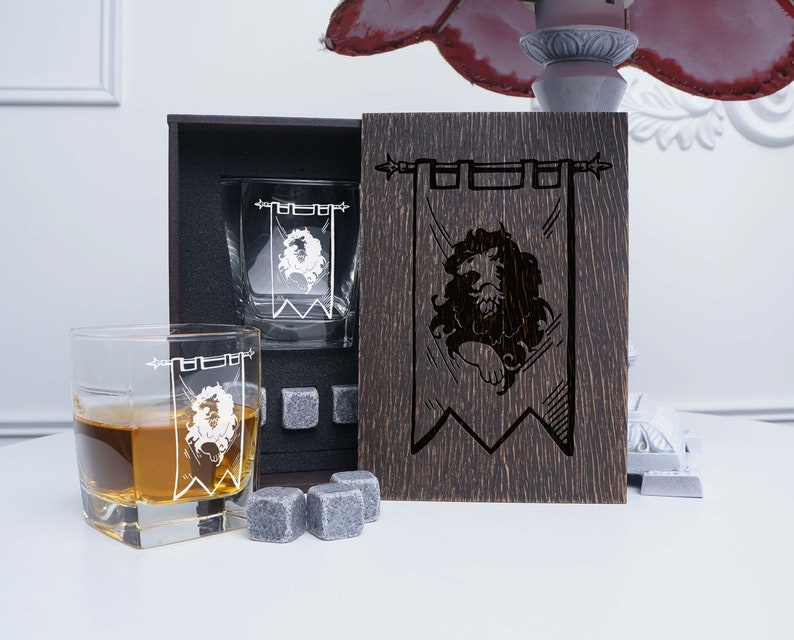 May include: Two clear glass whiskey tumblers with a white lion emblem on a banner design. The tumblers are in a wooden box with a matching wooden panel featuring the same lion emblem design.