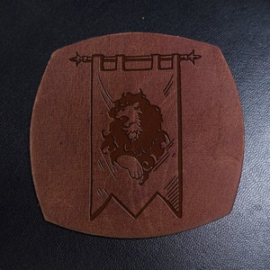 May include: A brown leather coaster with a raised design of a lion on a banner. The banner has a pointed top and bottom and is decorated with a simple border.