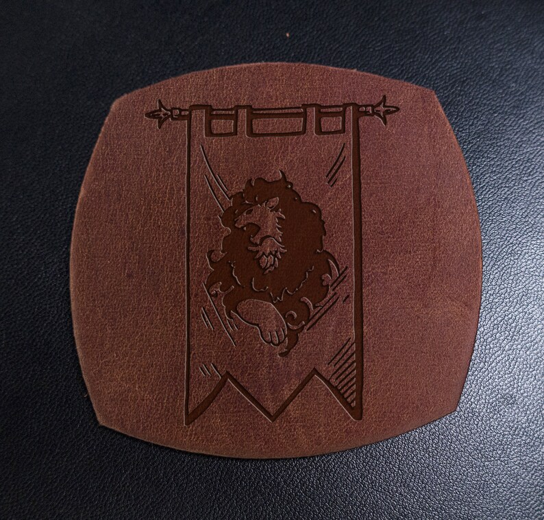 May include: A brown leather coaster with a raised design of a lion on a banner. The banner has a pointed top and bottom and is decorated with a simple border.