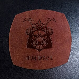May include: A square-shaped brown leather coaster with rounded edges featuring a dark embossed graphic of a samurai helmet and face mask. The name MICHAEL is embossed in a gothic-style font below the samurai illustration on the leather surface.