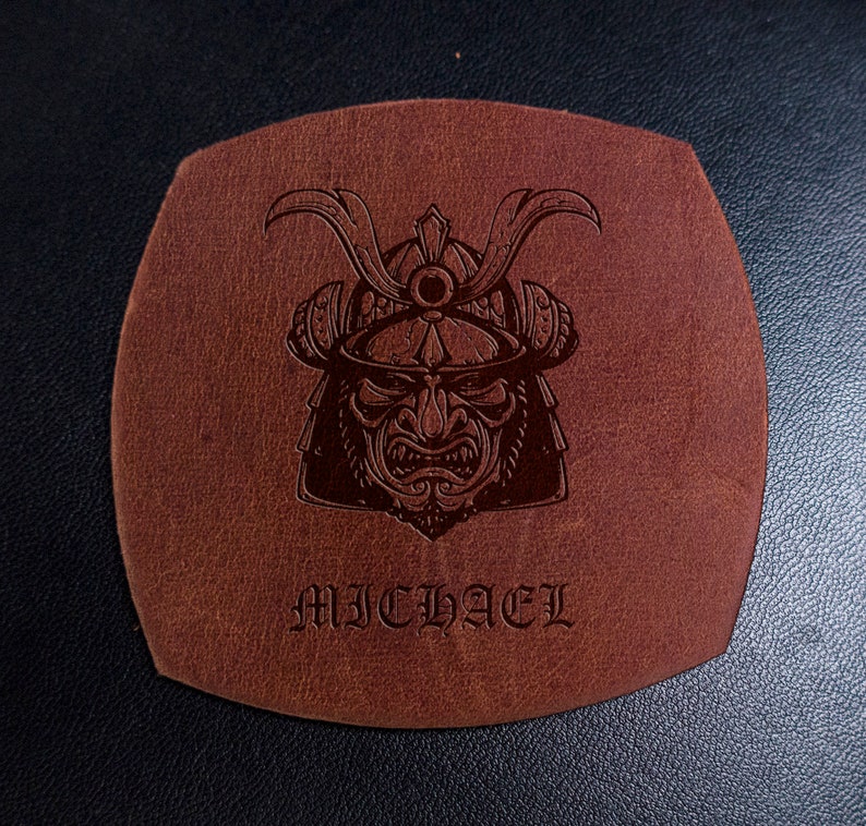 May include: A square-shaped brown leather coaster with rounded edges featuring a dark embossed graphic of a samurai helmet and face mask. The name MICHAEL is embossed in a gothic-style font below the samurai illustration on the leather surface.