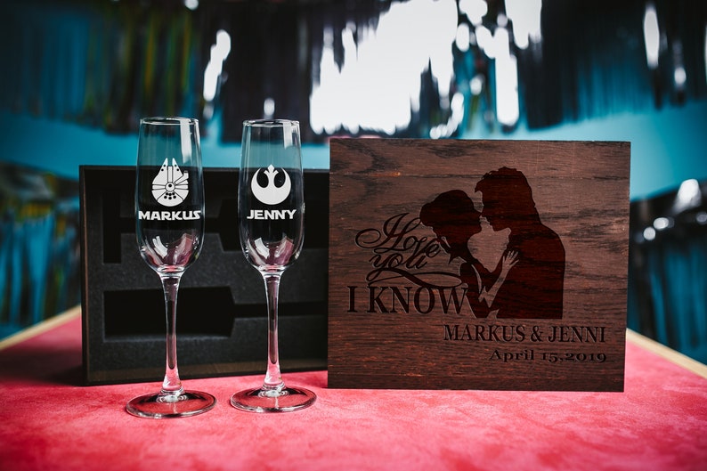May include: Two champagne flutes with Star Wars themed engravings, one with the Millennium Falcon and the other with the Rebel Alliance symbol. The flutes are in a black box with a wooden lid that says "I love you, I know" with a silhouette of Han Solo and Princess Leia. The lid also has the names "Markus & Jenni" and the date "April 15, 2019".