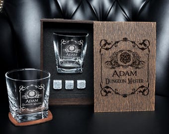 May include: A set of two clear glass tumblers, three grey stone ice cubes, and a dark wooden box. The glasses and the box lid are engraved with a decorative frame, a twenty-sided die icon, and the text Adam Dungeon Master.