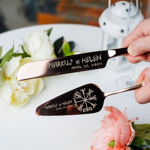 May include: A set of two rose gold cake knives with engraved details. The larger knife has the text "MARKUS & HELEN APRIL 25, 2024" and the smaller knife has a compass design and the same text.