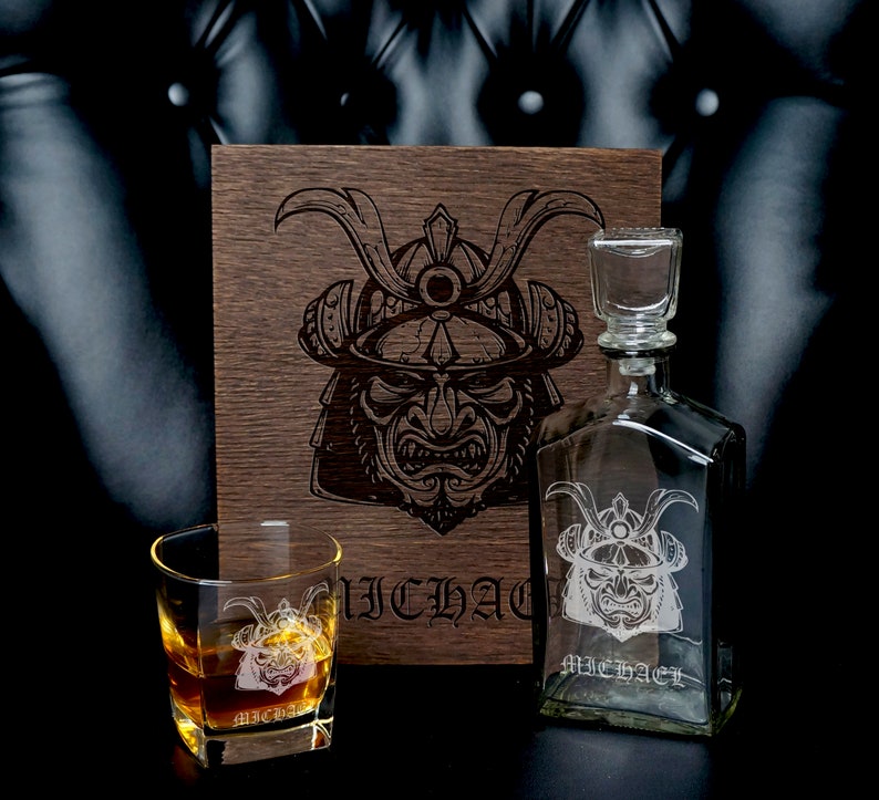 May include: A clear glass decanter and a matching whiskey glass, both etched with a samurai helmet design and the name MICHAEL. A dark wooden box stands behind them, featuring a large, detailed laser-engraved samurai helmet graphic.