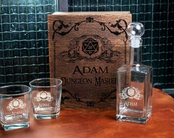 May include: A clear glass decanter with a matching stopper and two square whiskey glasses, all etched with a 20-sided die emblem and the name Adam. Behind them sits a rectangular wooden box engraved with the same emblem and the text Adam Dungeon Master.
