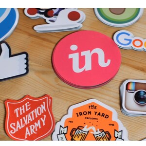 May include: A collection of colorful stickers featuring various logos, including Facebook, Instagram, Google, LinkedIn, and The Salvation Army. The stickers are arranged on a wooden surface.