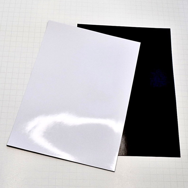 May include: Two sheets of paper, one white and one black, are lying on a white gridded surface. The white sheet is glossy and the black sheet is matte.