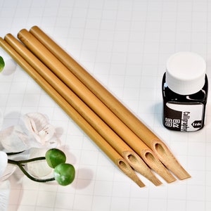Peut inclure: Quatre plumes de calligraphie en bambou avec un flacon d'encre de calligraphie noire. Le flacon d'encre a un bouchon blanc et une étiquette qui dit "Calligraphy Ink".