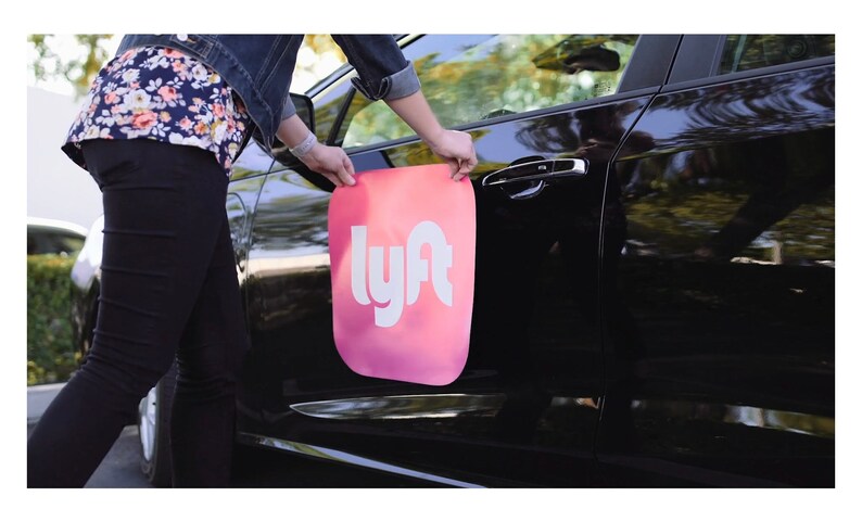 May include: A person places a pink and white Lyft logo magnet on the side of a black car.