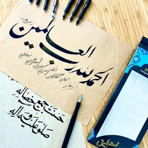 May include: A collection of calligraphy tools and Arabic script. The image shows black calligraphy pens, ink cartridges, and handwritten text on parchment paper. A box of calligraphy nibs is also present.