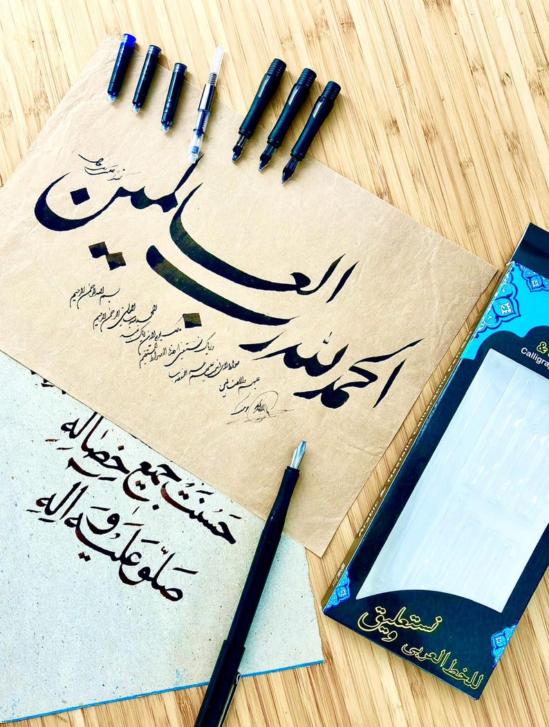 May include: A collection of calligraphy tools and Arabic script. The image shows black calligraphy pens, ink cartridges, and handwritten text on parchment paper. A box of calligraphy nibs is also present.