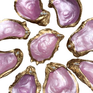 Op de afbeelding: Een verzameling van acht oesterschelpen met iriserende roze binnenkanten en metallic goudkleurige randen. Elke schelp is voorzien van kleine witte bolvormige parels aan de rand, wat een decoratieve set van schaaltjes vormt met een gladde afwerking.