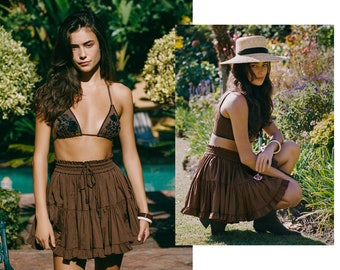 May include: A brown tiered skirt with a drawstring waist and a matching brown bikini top with floral embroidery. The model is wearing a straw hat and is sitting in a garden.