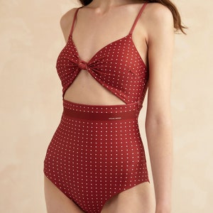 May include: A red one-piece swimsuit with white polka dots and a knotted detail at the bust. The swimsuit has a cutout at the waist and a black band with the text "VOGUE MODA" printed on it.