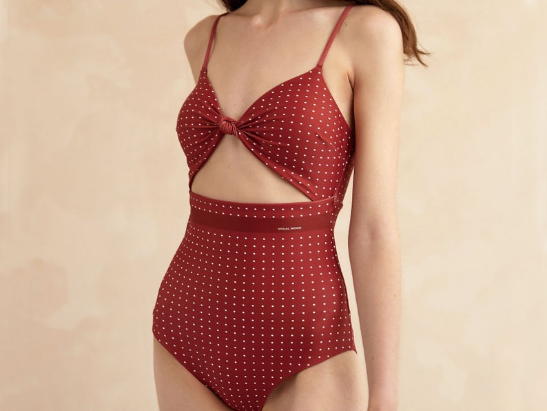 May include: A red one-piece swimsuit with white polka dots and a knotted detail at the bust. The swimsuit has a cutout at the waist and a black band with the text "VOGUE MODA" printed on it.