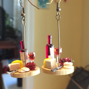 Puede incluir: Un par de pendientes con tablas de charcutería de vino y queso en miniatura. Cada pendiente presenta una pequeña botella de vino tinto, una copa de vino, una cuña de queso, un pequeño cuchillo y un racimo de uvas rojas.