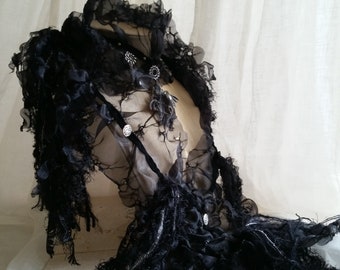 May include: A black sheer lace top with a ruffled collar and silver embellishments. The top is draped over a mannequin and has a flowing, ethereal look.