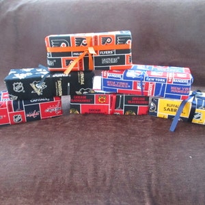 Puede incluir: Una colección de siete pequeñas cajas de regalo de tela con varios logotipos de equipos de la NHL, incluidos los Capitals, los Penguins, los Flyers, los Rangers, los Blackhawks y los Sabres. Las cajas están atadas con una cinta negra y azul.