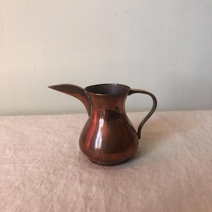 May include: A small copper pitcher with a flared spout and a handle. The pitcher has a hammered finish and a rounded body.