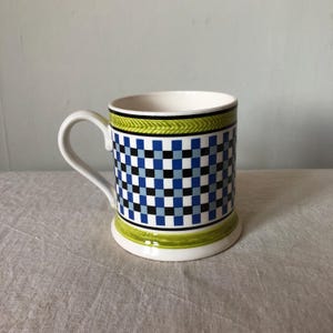 Könnte beinhalten: Eine Keramiktasse mit weißem Henkel und Innenraum. Die Außenseite weist ein kariertes Muster in blauen und schwarzen Quadraten auf, mit grünen und schwarzen Bändern oben und unten. Die Tasse ist etwa 10 cm hoch.
