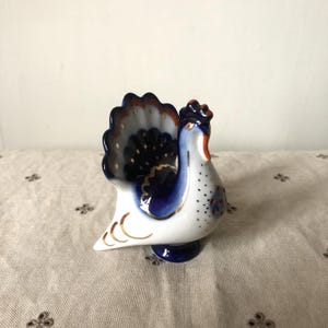 Pode incluir: Uma figurinha de porcelana branca de um pavão com detalhes azuis e dourados. O pavão tem as penas da cauda estendidas e está em pé sobre uma base.