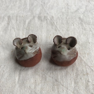 Peut inclure: Deux petites figurines de souris en céramique. Chaque souris a une base arrondie brun-rougeâtre et une tête avec un visage blanc et gris, de petites oreilles et des moustaches. Les figurines sont faites à la main et ont un aspect rustique.
