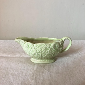 May include: A light green ceramic gravy boat featuring a raised, embossed leaf pattern across its exterior. The vessel has a scalloped rim, a curved pouring spout, and a single looped handle, all finished in a glossy, pale green glaze.