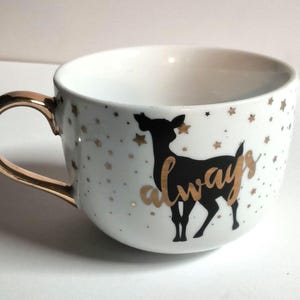 May include: A white ceramic mug featuring a black silhouette of a deer and the word always written in gold script. The mug is decorated with small gold stars and has a metallic gold handle. The interior of the mug is plain white ceramic.