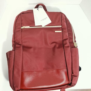 May include: A burgundy backpack featuring a smooth fabric upper section and a textured faux leather base. It includes a horizontal front zipper pocket with gold-tone hardware, side pockets, and a white product tag with the text LOVEVOOK.