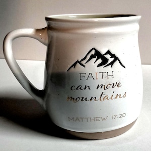 May include: A white ceramic mug with a speckled finish and a curved handle. The front features a black mountain graphic above the metallic copper text "FAITH can move mountains" and the reference "MATTHEW 17:20" in a simple sans-serif font.