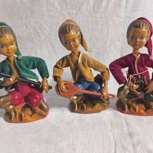 May include: Three figurines of musicians in seated poses. The left figure wears a green jacket and red pants, holding a black flute. The center figure wears a tan jacket and blue pants, holding a lute. The right figure wears a red jacket and green pants, holding a lyre.