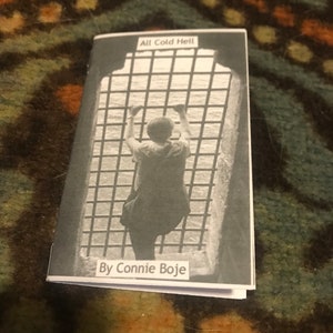 Puede incluir: Una fotografía en blanco y negro de una persona que alcanza una ventana enrejada. El texto "All Cold Hell" está en la parte superior de la imagen y "By Connie Boje" en la parte inferior.