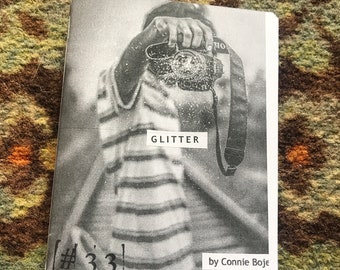 Könnte beinhalten: Ein Schwarzweißfoto einer Person, die eine Kamera hält. Das Foto ist auf einem weißen Papierheft mit dem Titel "GLITTER #33" von Connie Boje gedruckt.