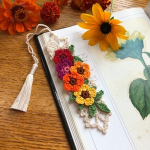 May include: A crocheted bookmark with a tassel, featuring a cascade of colourful flowers in shades of red, orange, and yellow. The bookmark is resting on a book with a botanical illustration of a sunflower.