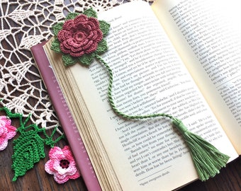 May include: A crocheted bookmark with a pink flower and green leaves. The bookmark has a green tassel at the bottom. The bookmark is resting on a book with the text "Spiny evergreen shrub."