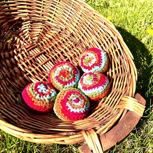 May include: A large, round, brown woven wicker basket containing five small, spherical, crocheted balls. Each ball features a vibrant, multi-colored spiral pattern in shades of red, green, brown, and light blue, crafted with a textured yarn finish.