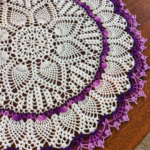 May include: A white crocheted doily with a purple and pink ombre border. The doily is circular and has a delicate lace pattern.