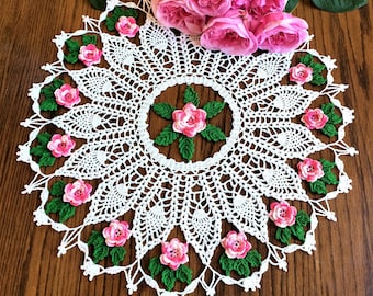 May include: White crocheted doily with a floral design. The doily is decorated with pink and green crocheted flowers. The doily is on a wooden surface.